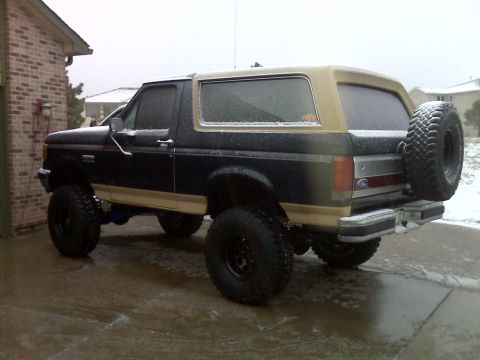 1990 Ford Bronco - The rear passenger side of a lifted, dark blue and tan Ford Bronco is shown parked on a wet, snowy driveway. A large spare tire is mounted…