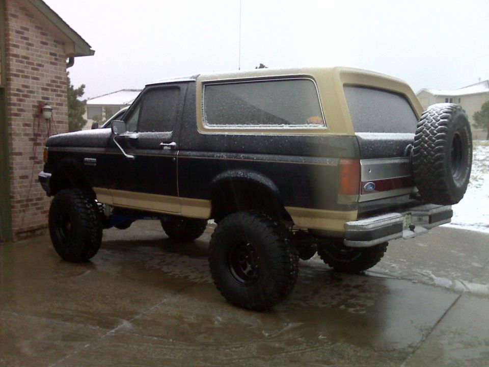 Lifted 1990 Ford Bronco with spare tire on rear door, parked on snowy driveway.