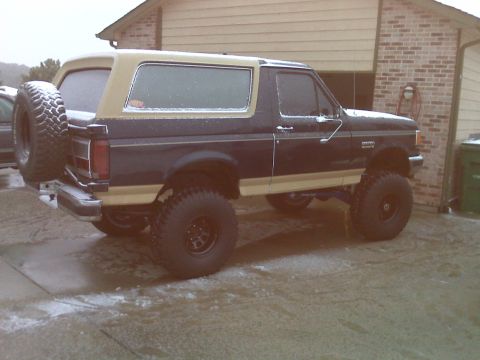 1990 Ford Bronco - A dark blue and tan Ford Bronco is parked on a wet, snowy driveway in front of a brick house. The vehicle has large off-road tires and a sp…