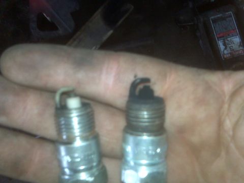 1995 Ford Bronco - A hand holds two spark plugs, one with a clean electrode and the other with a heavily fouled electrode covered in black carbon deposits.