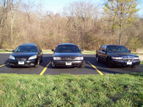 1999 Mercury Grand Marquis - Three black sedans are parked in a lot with yellow lines. The car on the right is a 1999 Mercury Grand Marquis.