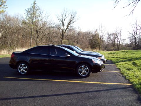 1999 Mercury Grand Marquis - Two black sedans are parked side-by-side in a paved lot with yellow parking lines. The car in the foreground is a Pontiac G6 with tinted wi…