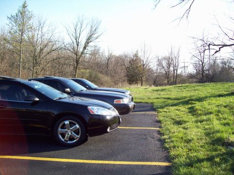 1999 Mercury Grand Marquis - Three black sedans are parked in a row on asphalt with yellow parking lines. The foreground car is a Pontiac G6, and the two cars behind it…