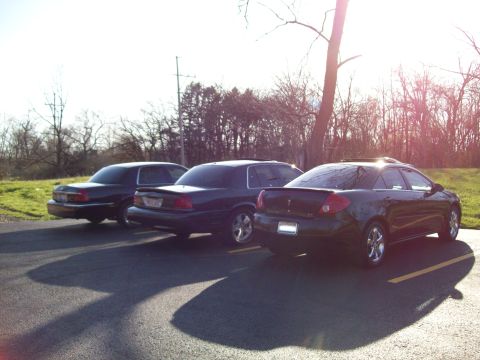1999 Mercury Grand Marquis - Three dark-colored sedans are parked in a lot with a grassy area and trees in the background. The sun is shining brightly, casting long sha…