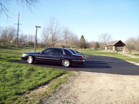 1999 Mercury Grand Marquis - A dark blue 1999 Mercury Grand Marquis sedan is parked on an asphalt lot next to a grassy area. A small wooden pavilion is visible in the b…