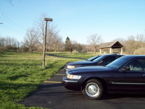 1999 Mercury Grand Marquis - A dark blue sedan parked on an asphalt lot next to a grassy area. A picnic shelter with a wooden roof is visible in the background.