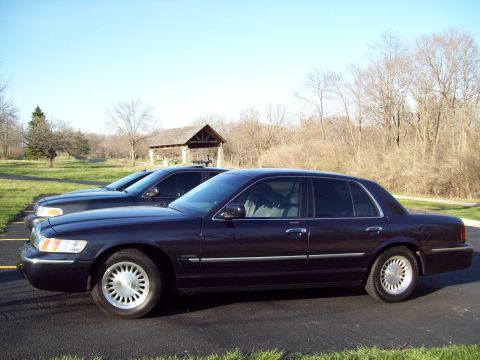 1999 Mercury Grand Marquis - A dark colored sedan is parked on asphalt with grass and trees in the background. Another sedan is parked behind and to the left of the mai…