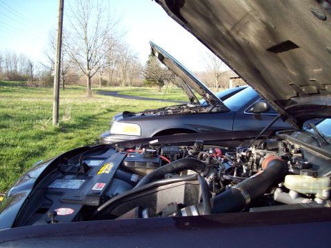 1999 Mercury Grand Marquis - The open hoods of two dark-colored sedans are shown, with the engine bay of the foreground vehicle prominently displayed. The background sh…