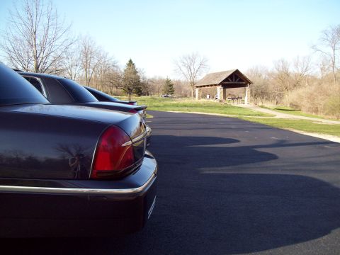 1999 Mercury Grand Marquis - The rear quarter panel and taillight of a dark colored sedan are visible in the foreground, parked on asphalt. A picnic shelter is visible…