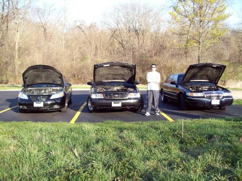 1999 Mercury Grand Marquis - A person stands between two black sedans with their hoods open, parked on asphalt with yellow lines. A third black sedan is parked to the l…