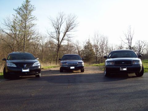 1999 Mercury Grand Marquis - Three black sedans are parked on asphalt in front of a wooded area. The car on the right is a Ford Crown Victoria, while the car on the lef…