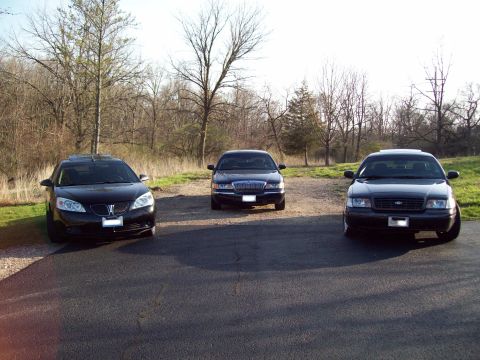 1999 Mercury Grand Marquis - Three dark-colored sedans are parked on a paved surface with a wooded area in the background. The car on the left is a black Pontiac G6, th…