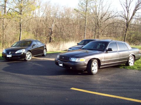 1999 Mercury Grand Marquis - Two sedans are parked on an asphalt surface with a yellow line. A dark-colored sedan is in the foreground, and a black sedan is parked to i…
