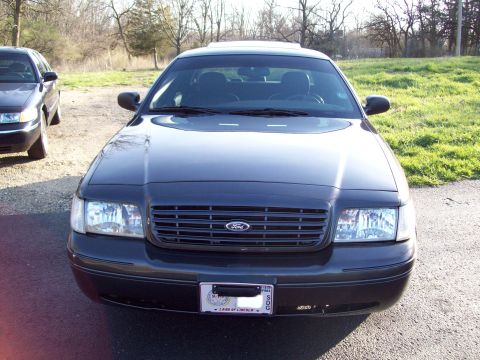 1999 Mercury Grand Marquis - The front view of a dark-colored sedan, likely a Ford Crown Victoria, is shown parked outdoors on a gravel and asphalt surface with a grass…