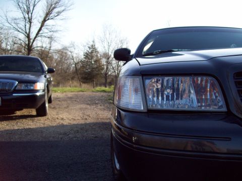 1999 Mercury Grand Marquis - A close-up, low-angle view of the front right headlight and grille of a dark-colored sedan parked outdoors. Another dark-colored sedan is v…
