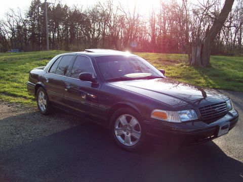 1999 Mercury Grand Marquis - A dark colored, four-door sedan is parked on an asphalt driveway with a gravel shoulder. The car is angled towards the camera, showing its…