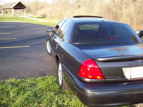 1999 Mercury Grand Marquis - The rear passenger side of a dark gray sedan is parked on a grassy area next to a paved parking lot. The car has tinted windows and a spoil…