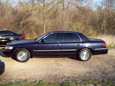 1999 Mercury Grand Marquis - The side view of a dark blue four-door sedan parked on a gravel surface with dry brush and trees in the background. A second dark-colored v…