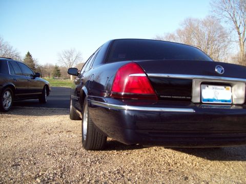 1999 Mercury Grand Marquis - The rear passenger side of a dark blue Mercury Grand Marquis is shown parked on a gravel surface. The vehicle has an Illinois license plate…