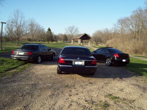 1999 Mercury Grand Marquis - Three dark sedans parked on a gravel lot at a park with a stone-and-timber picnic shelter and bare early-spring trees behind them. Center i…