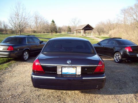 1999 Mercury Grand Marquis - The rear view of a dark blue Mercury Grand Marquis is parked on a gravel driveway in front of a picnic shelter. Another dark sedan is parke…