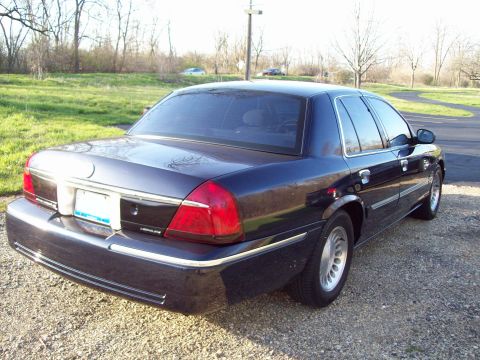 1999 Mercury Grand Marquis - The rear passenger side of a dark blue sedan is shown parked on a gravel surface. The car has tinted windows and chrome trim along the bott…