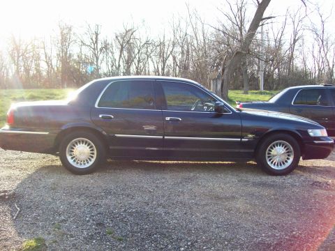 1999 Mercury Grand Marquis - A dark blue Mercury Grand Marquis sedan is parked on a gravel surface with trees and grass in the background. The sun is shining brightly,…