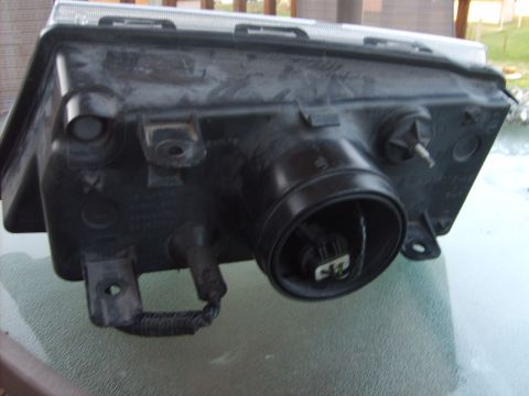 1993 Ford Crown Victoria - The rear housing of a black automotive headlight assembly is shown from a low angle, resting on a textured surface. The assembly features a…