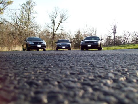 1999 Mercury Grand Marquis - Three sedans are parked on the side of a road with trees in the background. The closest car is a black sedan, followed by a dark blue sedan…