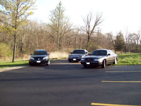 1999 Mercury Grand Marquis - Three sedans are parked in a paved lot with a wooded area in the background. The closest vehicle is a dark sedan, followed by a dark sedan…