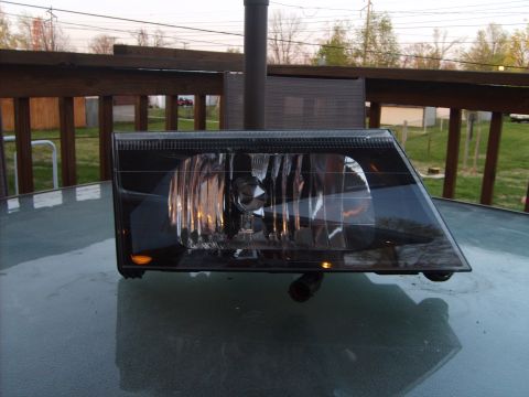 1993 Ford Crown Victoria - A black aftermarket headlight assembly, likely for a 1993 Ford Crown Victoria, rests on a glass table outdoors. The headlight features a cl…
