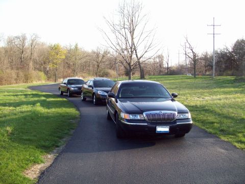 1999 Mercury Grand Marquis - A black 1999 Mercury Grand Marquis is parked on a paved road next to a grassy area with trees in the background. The car is facing towards…