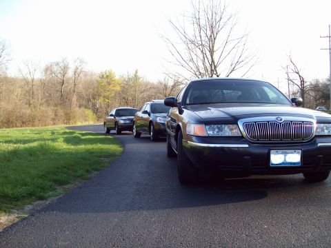 1999 Mercury Grand Marquis - A black Mercury Grand Marquis is parked on the side of a paved road, with other cars behind it. The car is facing the camera at a low angle…