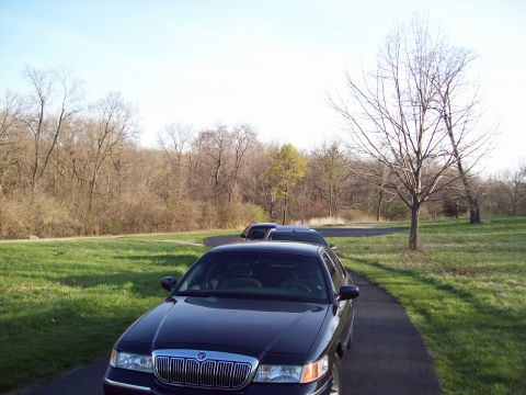1999 Mercury Grand Marquis - A dark blue 1999 Mercury Grand Marquis is parked on a paved path next to a grassy area. The car is viewed from the front 3/4 angle, showing…