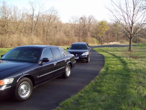 1999 Mercury Grand Marquis - Two dark-colored sedans are parked on a paved driveway surrounded by grass and trees. The car in the foreground is a dark blue sedan, and t…