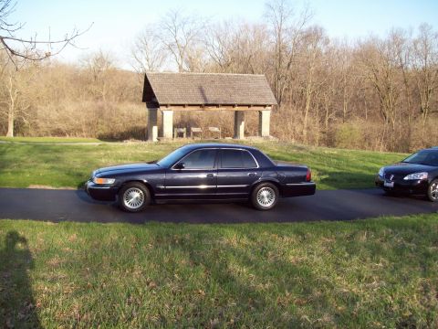 1999 Mercury Grand Marquis - dark blue Mercury full-size sedan parked on pavement near a stone picnic shelter and bare trees.