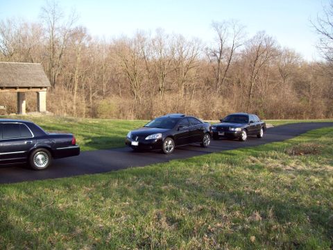 1999 Mercury Grand Marquis - Three black sedans are parked on a paved road next to a grassy area with trees in the background. The car on the left is a dark sedan, and…