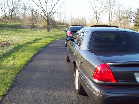 1999 Mercury Grand Marquis - A dark gray sedan is parked on a paved driveway next to a grassy area. The rear of the car is visible, showing its taillight and a spoiler.