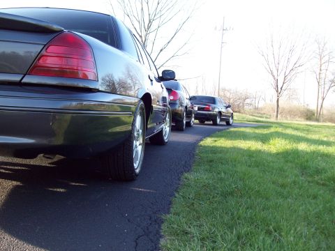 1999 Mercury Grand Marquis - A dark gray sedan is parked on an asphalt driveway next to a grassy area. The rear of the car is in the foreground, with two other sedans v…