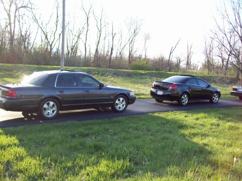 1999 Mercury Grand Marquis - A dark gray sedan is parked on an asphalt road next to a black sedan. The vehicles are positioned in a grassy area with trees in the backgr…