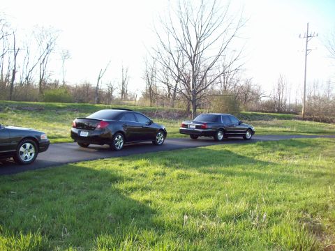 1999 Mercury Grand Marquis - Three dark sedans are parked on a paved surface next to a grassy area. The car closest to the viewer is partially visible on the left, whil…