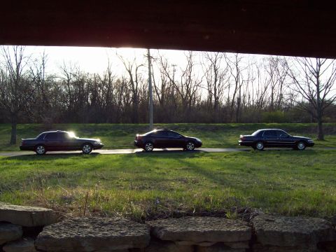 1999 Mercury Grand Marquis - Three dark sedans are parked in a line on a paved surface next to a grassy area. The sun is setting behind the trees, casting a warm glow o…