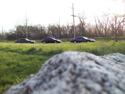 1999 Mercury Grand Marquis - A black sedan is parked on the side of a grassy road with trees in the background. The car is viewed from a low angle, with a large rock in…