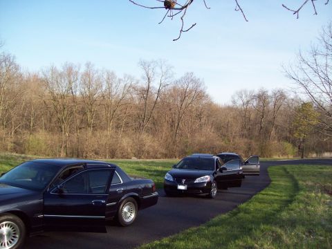 1999 Mercury Grand Marquis - Two dark-colored sedans are parked on a paved road next to a grassy area and a line of bare trees under a clear blue sky. The sedan in the…