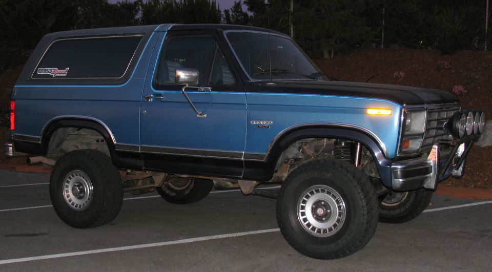 Blue 1984 Ford Bronco with black roof and off-road lights, parked outdoors.