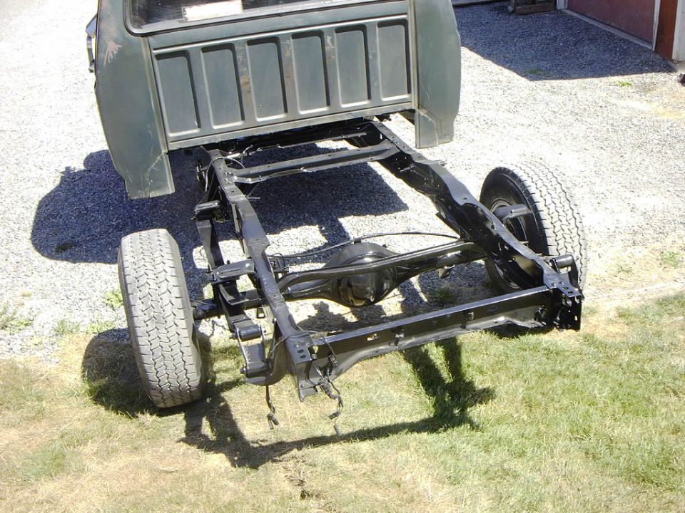 Rear view of green pickup with bed removed, showing black-painted frame and rear axle on gravel