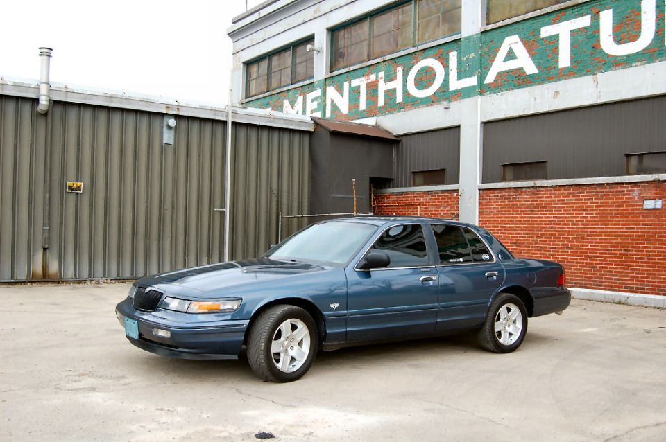 Blue-grey Mercury Grand Marquis parked outside an old brick Mentholatum factory building