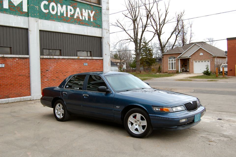 Blue 1990s Mercury Grand Marquis sedan on alloy wheels beside an old brick building