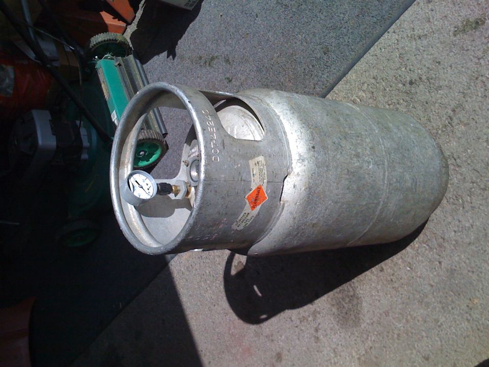 Propane tank with pressure gauge next to a lawnmower.