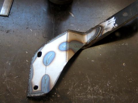 1996 Ford Bronco - A fabricated steel bracket lying on a scratched shop table, welded from a shaped plate with two bolt holes and a long flat arm, showing blu…